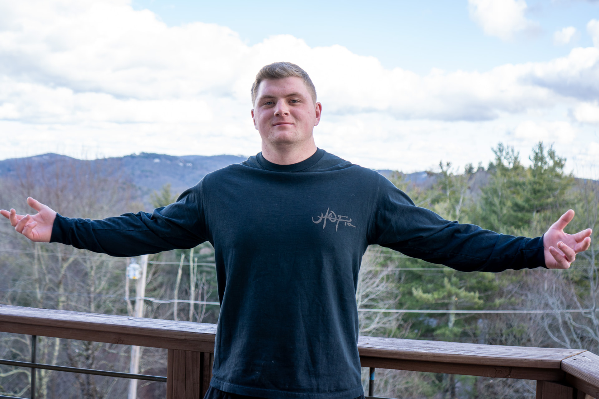 Nolan Blakeman with arms spread wide on a wooden deck with mountains behind him